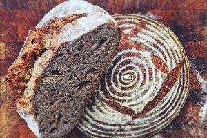 A loaf of rustic sourdough bread on a wooden board, partially sliced to reveal its dense, airy crumb. The unsliced portion displays a decorative spiral and leaf pattern dusted with flour on the crust.